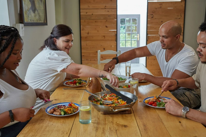 Four adults passing a pan of food around a sunlit wooden dining table