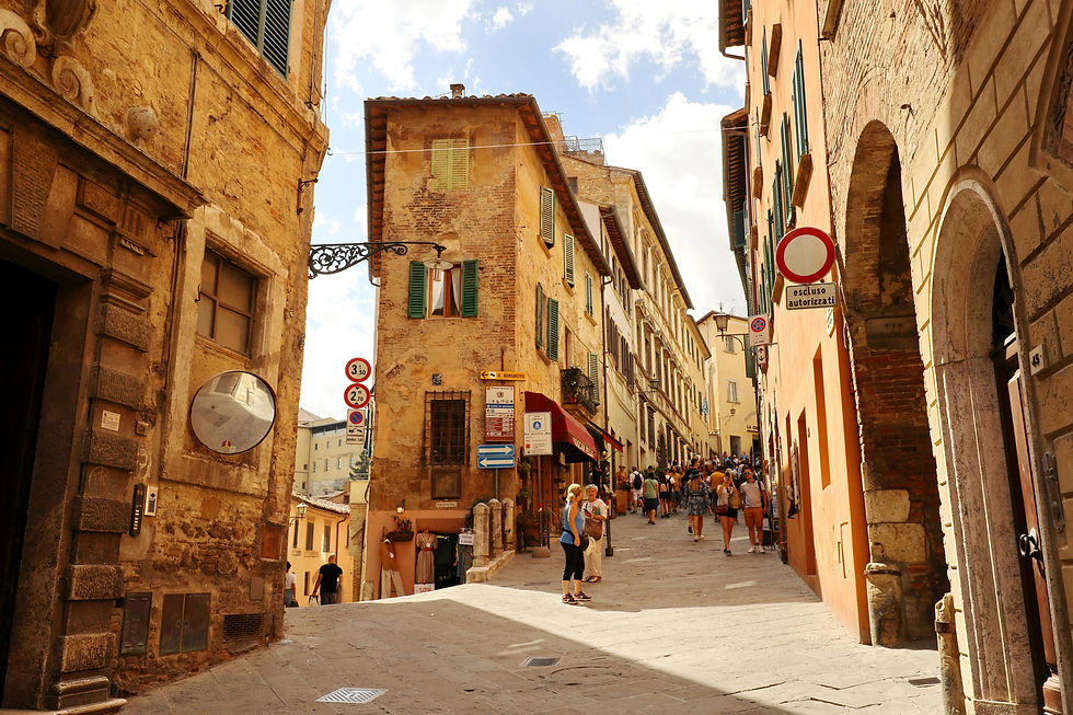 People walk up a sunlit, narrow cobblestone street flanked by historic yellow and orange buildings with green shutters and signs in Italian. Montepulciano, Tuscany