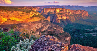 Cânion com penhascos alaranjados ao pôr do sol, vegetação verde no vale e céu rosado; paisagem majestosa e calma.