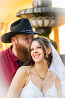 A groom with a black cowboy hat and dark red shirt leans into to give his glowing bride in a white a kiss on the head.