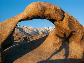 a dancers shadow is cast on the orange granite rock arch lit by the morning sun. The arch is a window to the distant jagged mountain peaks