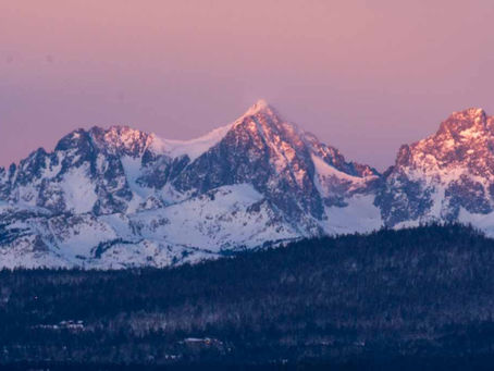 Ritter and Banner in the Eastern Sierras are snowy mountain peaks glow pink at sunset above dark forested hills under a pastel sky.
