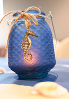 A blue dimpled jar is lit by a candle. A gold sea horse, tied by a cord situates the seahorse in front of the glow from the candle.