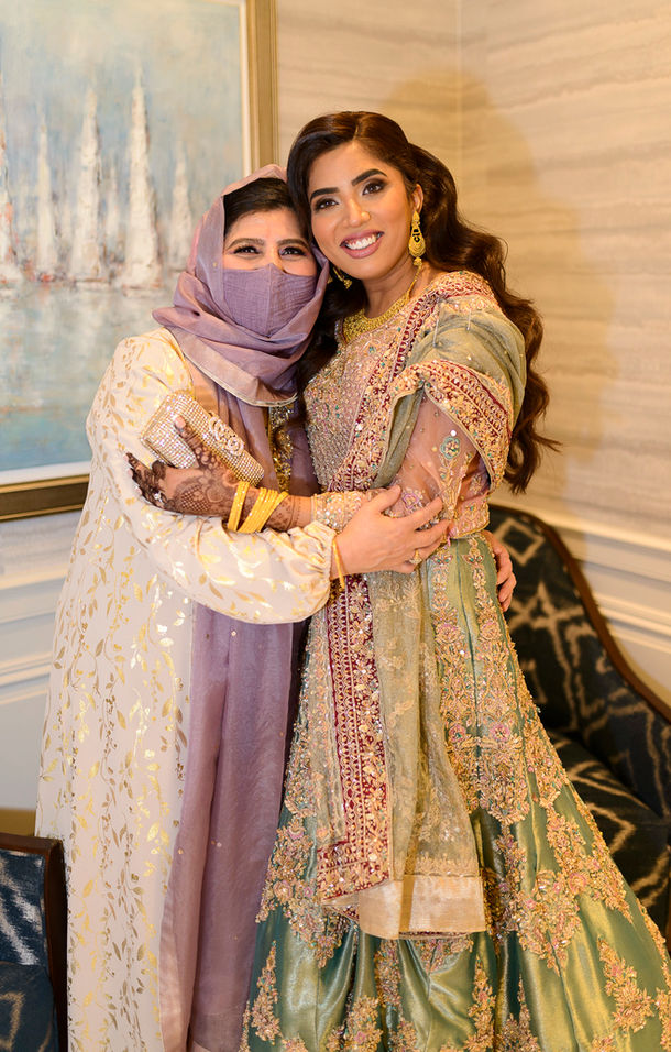 A joyful portrait of the bride embracing the groom's mother at a Pakistani wedding. The bride wears a stunning green and gold traditional outfit with intricate embroidery and maroon accents, paired with gold jewelry. The groom's mother is dressed elegantly in a lavender and gold ensemble with a matching scarf and holds a beaded clutch. Both women share warm smiles, showcasing a moment of love and family connection against a softly lit, elegant indoor background.