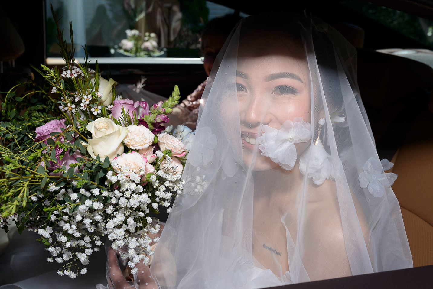 A bride sits in a car, photographed through the window, her face softly framed by a delicate white veil adorned with floral accents. She smiles warmly, holding a bouquet of white, pink, and purple flowers with lush greenery and baby's breath. The scene captures a moment of serene joy at a New England wedding.