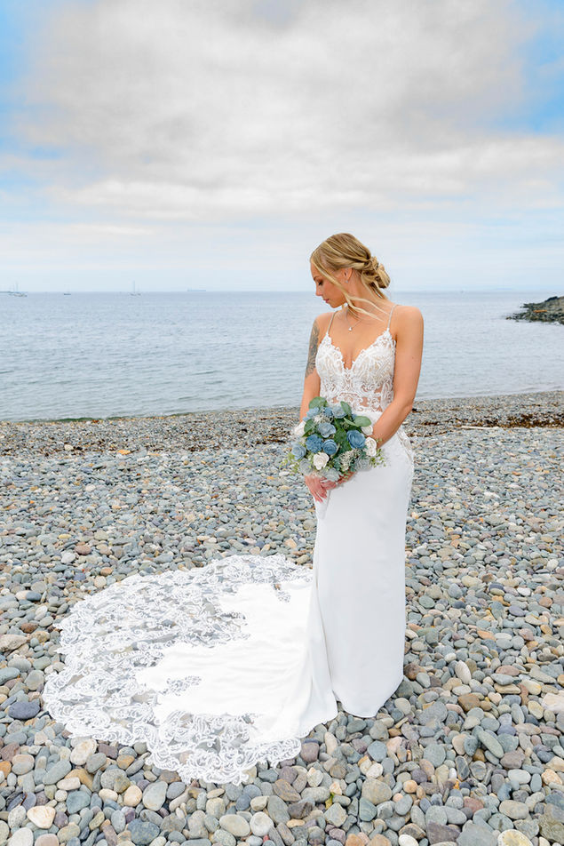 Elegant bride standing alone on a pebble beach, gazing downward with a serene expression. She holds a bouquet of white and blue flowers, matching her intricate lace-detailed wedding dress that flows into a detailed train on the rocky ground. The overcast sky and calm sea provide a peaceful backdrop to her reflective moment.