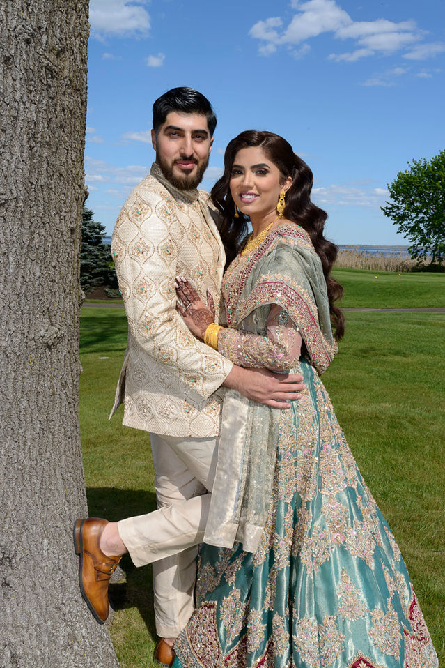 A joyous Pakistani wedding portrait featuring the bride and groom embracing beside a large tree. The groom wears a cream-colored sherwani adorned with gold and green embroidery, paired with beige trousers and brown leather shoes. The bride is dressed in a stunning sage green lehenga heavily embellished with intricate gold and pink embroidery, paired with traditional gold jewelry and her henna-adorned hands resting on the groom. The couple stands on a lush green lawn under a vibrant blue sky with scattered white clouds, creating a picturesque backdrop.