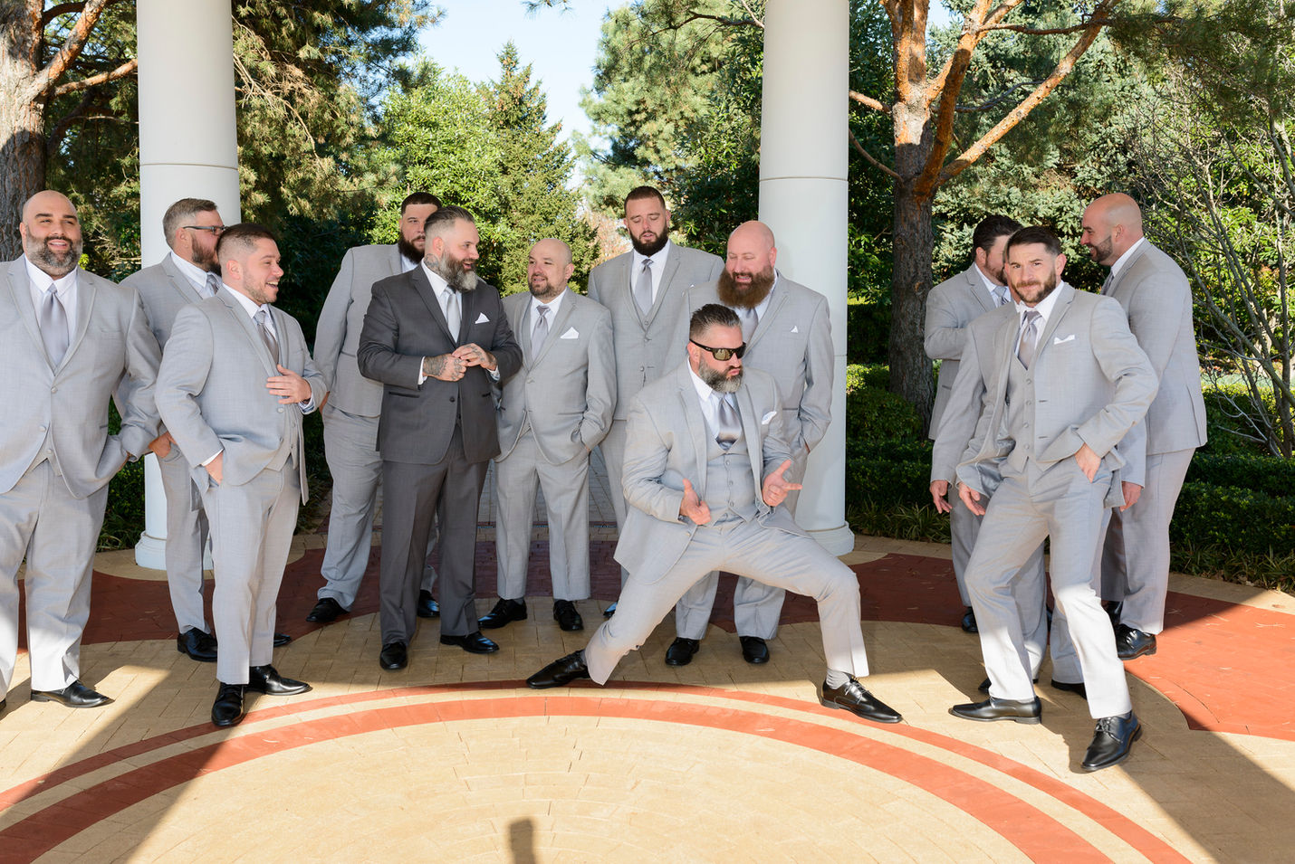 A group of groomsmen and the groom gather for a lively and humorous moment on the wedding day. The groomsmen, dressed in matching light gray suits with ties, surround the groom, who wears a dark gray suit. One groomsman strikes a playful pose in the foreground, adding energy and fun to the scene. They are standing under a columned structure, with greenery and trees in the background. The photo captures camaraderie and lighthearted celebration.