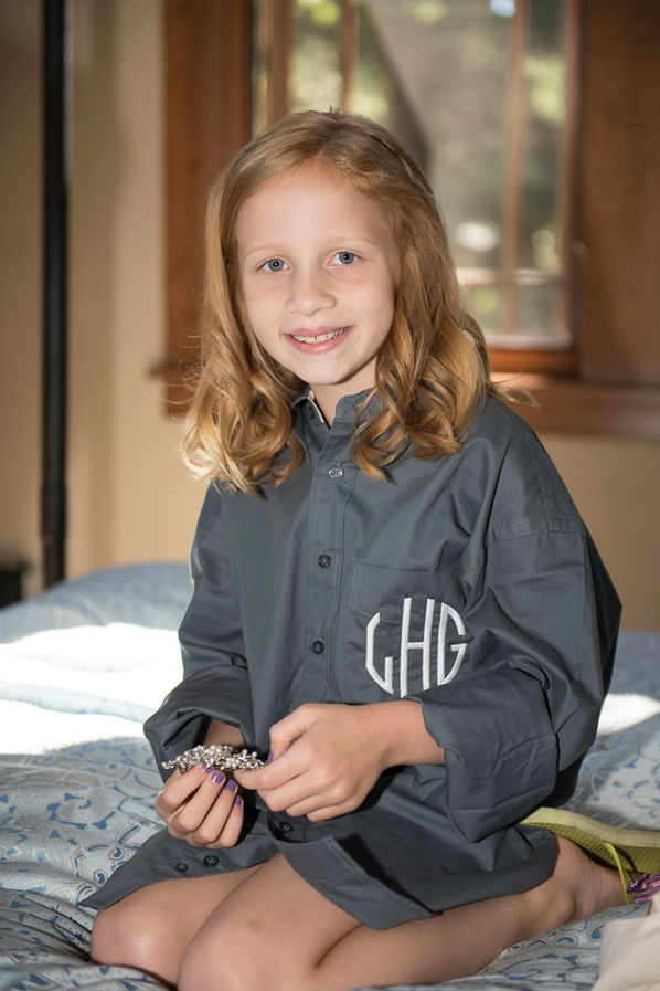 A young flower girl sits on a bed in the getting-ready room, wearing an oversized gray monogrammed shirt with the initials 'GHG.' She smiles sweetly while holding a sparkling bracelet, her strawberry-blonde hair styled in soft curls, illuminated by natural light streaming through the window behind her.