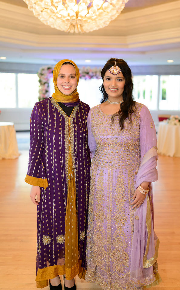 Two guests pose for a photograph at a Pakistani wedding celebration. One guest wears a vibrant purple and gold traditional outfit with intricate detailing and a matching golden hijab. The other guest is dressed in a pastel purple traditional outfit adorned with delicate golden embroidery and sparkling embellishments, paired with matching jewelry. Both are smiling warmly, standing under an elegant chandelier in a beautifully decorated reception hall.