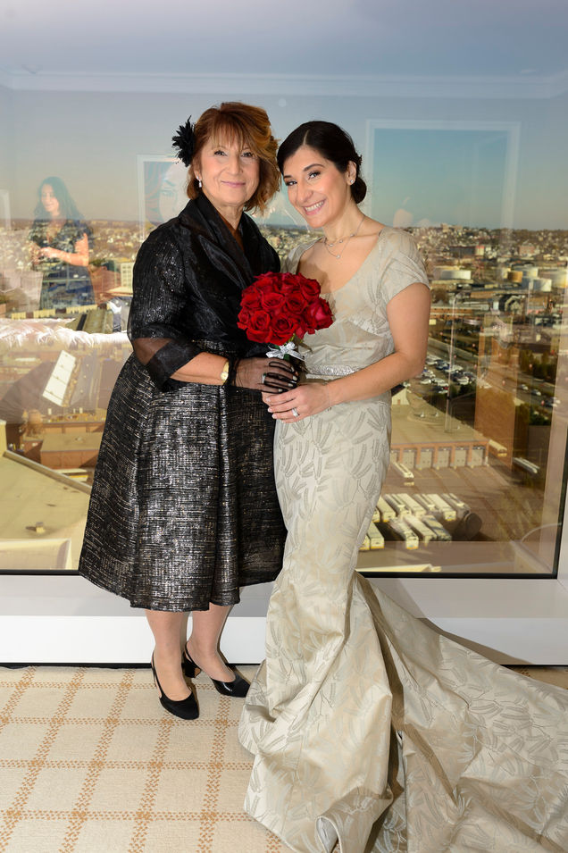 The bride and her mother pose together in the ready room, creating a beautiful wedding portrait. The bride wears an elegant light-colored gown with a flowing train and holds a bouquet of vibrant red roses. Her mother, dressed in a shimmering black dress with sheer gloves, stands proudly beside her. A large window in the background reveals a panoramic view of the city, adding depth and brightness to the image.