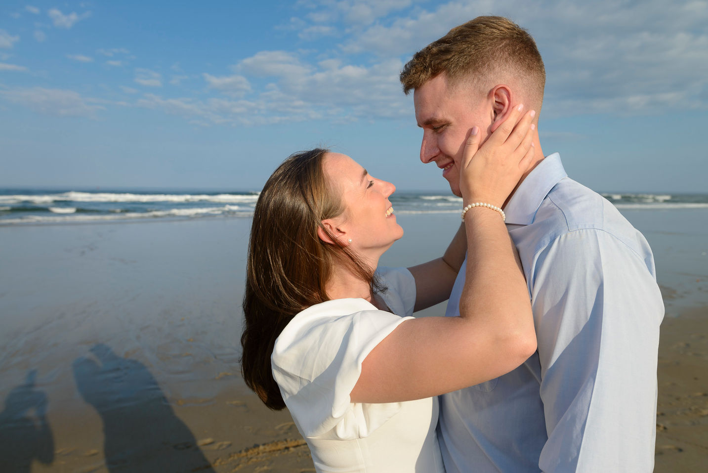 A romantic moment captured on the beach during an engagement photo session, with the woman gently holding the man's face, both smiling lovingly at each other against the backdrop of soft ocean waves and a clear blue sky.