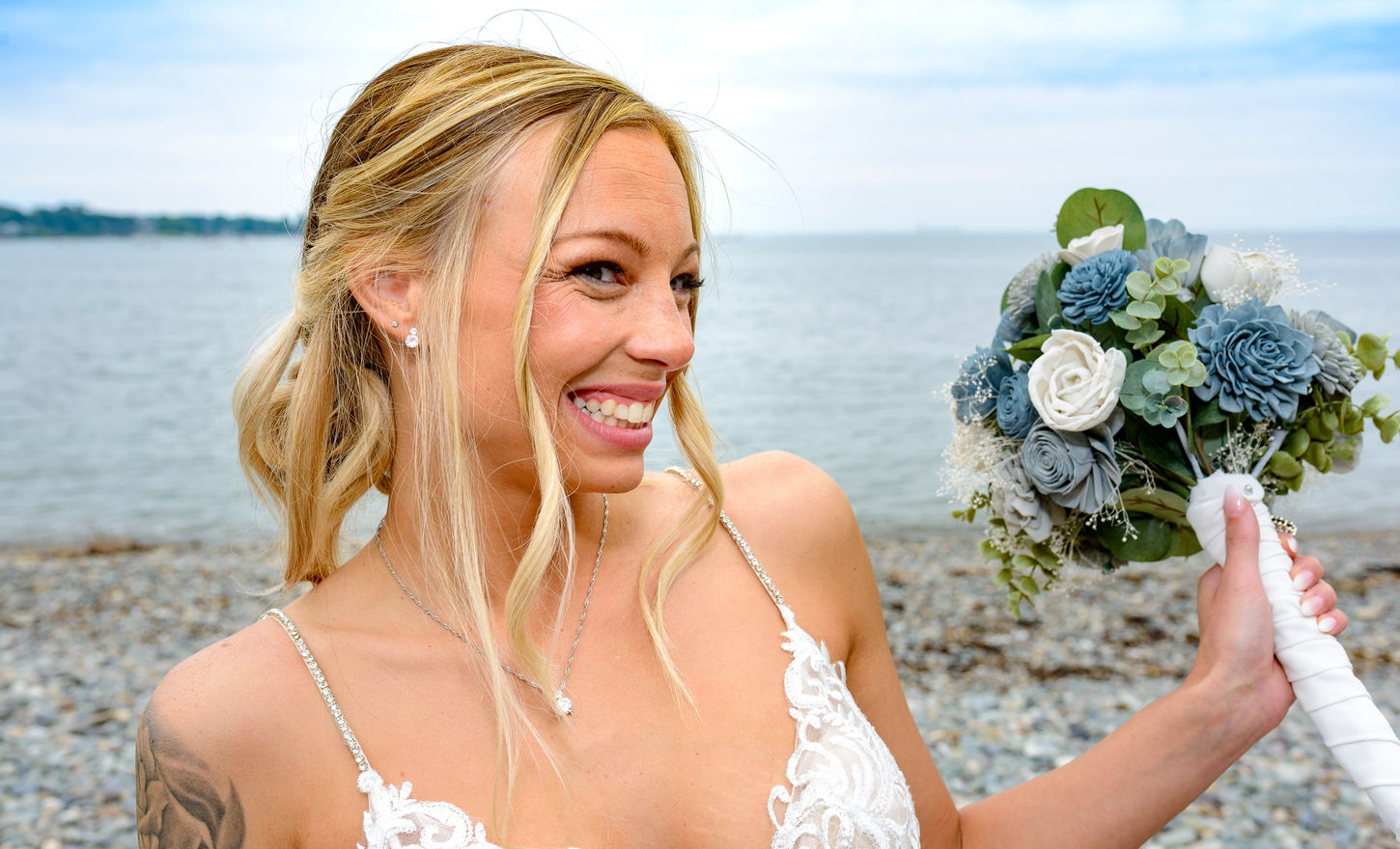 Close-up of a radiant bride at her seaside wedding, smiling broadly as she holds a bouquet of blue and white flowers. Her elegant lace wedding gown, detailed with delicate beading on the straps, complements her joyful expression. The ocean and a pebbled shore form a serene backdrop, enhancing the bride's natural beauty and happiness on her special day. Wedding portrait photographer Boston and New England.