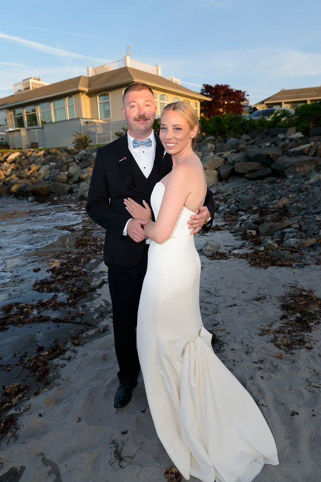 Bride and groom pose affectionately on a beach at sunset. The bride, clad in a sleek white wedding gown, smiles gently while wrapped in the groom's arms. The groom, in a classic black tuxedo with a white bow tie, looks towards the camera with a subtle smile. Behind them, the soft glow of the setting sun illuminates a seaside house and a rocky shore, enhancing the romantic ambiance of the scene.