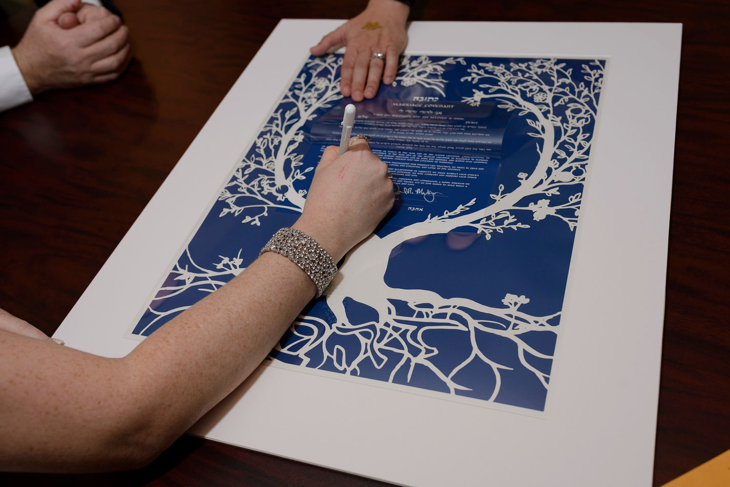 Close-up of the bride's hand signing a beautifully designed Ketubah, a traditional Jewish marriage contract, featuring an intricate white tree design on a deep blue background.