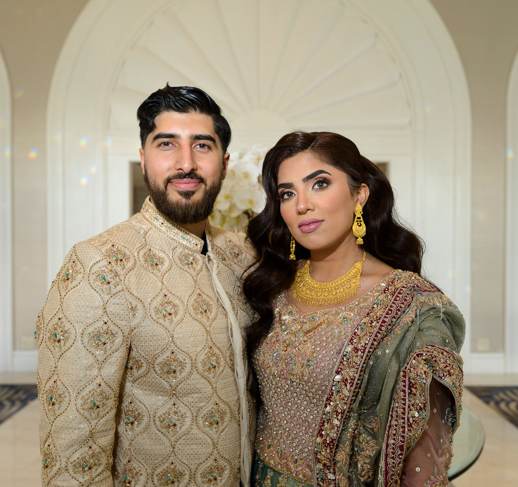 A close-up portrait of the bride and groom during a Pakistani wedding celebration. The groom is dressed in an intricately embroidered cream sherwani, and the bride wears a stunning teal and maroon lehenga adorned with gold accents. Her look is completed with elegant gold jewelry, including statement earrings and a necklace. Both look directly at the camera, exuding confidence and grace, with a beautifully ornate backdrop enhancing the setting.