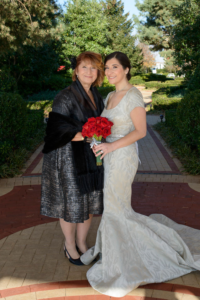 A wedding photograph featuring the bride and her mother. The bride is wearing a stunning ivory gown with a long, elegant train, holding a bouquet of vibrant red roses. Her mother is dressed in a shimmering black dress with a black shawl, standing proudly beside her daughter. The background showcases a beautifully landscaped garden with lush greenery, creating a serene and memorable setting for the special day.