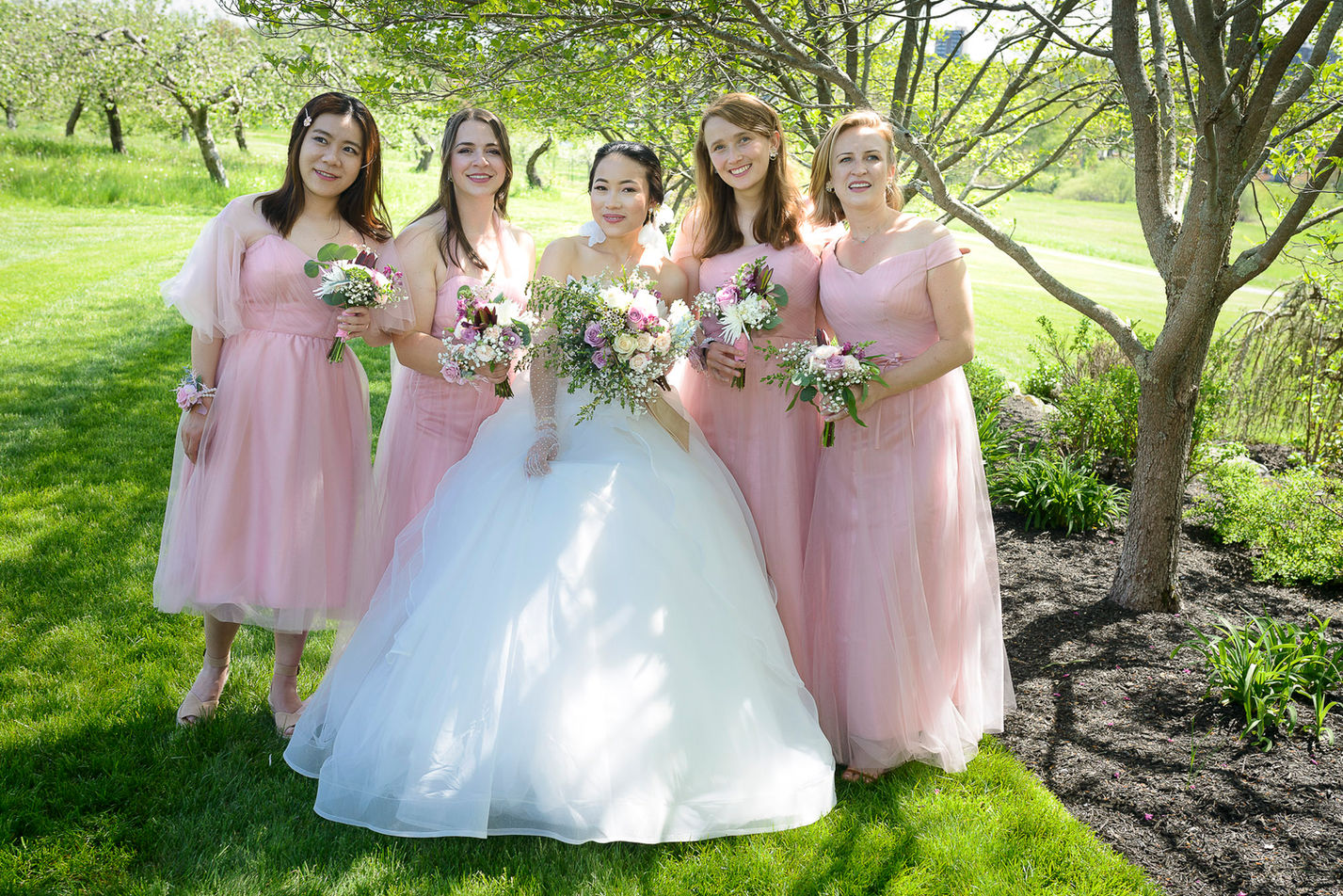 Bride standing with her bridesmaids in an outdoor apple blossom orchard setting. The bride wears a white gown and holds a pastel bouquet, while the bridesmaids are dressed in matching blush pink dresses, each holding smaller bouquets of roses and greenery. The group smiles warmly under the shade of leafy trees, with a sunny landscape in the background."