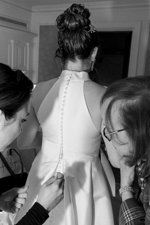 Black and white photo of a bride from behind as two women assist her with fastening the delicate buttons on the back of her elegant wedding gown. Her hair is styled in an intricate updo adorned with pearls, and the scene captures the care and detail of the final wedding preparations.
