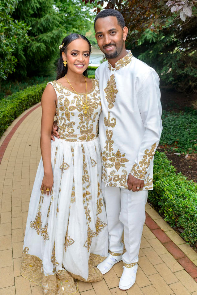 Bride and groom standing together on garden path in white and gold outfits.
