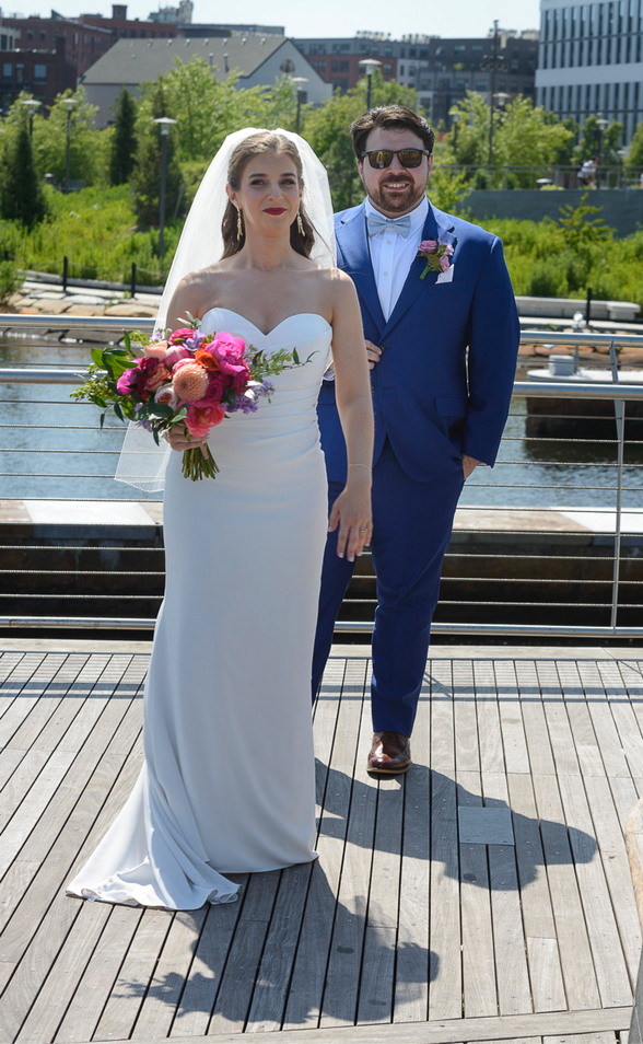 A stunning wedding photograph featuring the bride and groom on a wooden boardwalk near the water. The bride is wearing a strapless white gown with a long veil, holding a vibrant bouquet of colorful flowers, while the groom stands behind her in a sharp blue suit with a bow tie, sporting sunglasses. The backdrop includes lush greenery, modern architecture, and a calm waterfront, creating a picturesque outdoor scene on a sunny day.