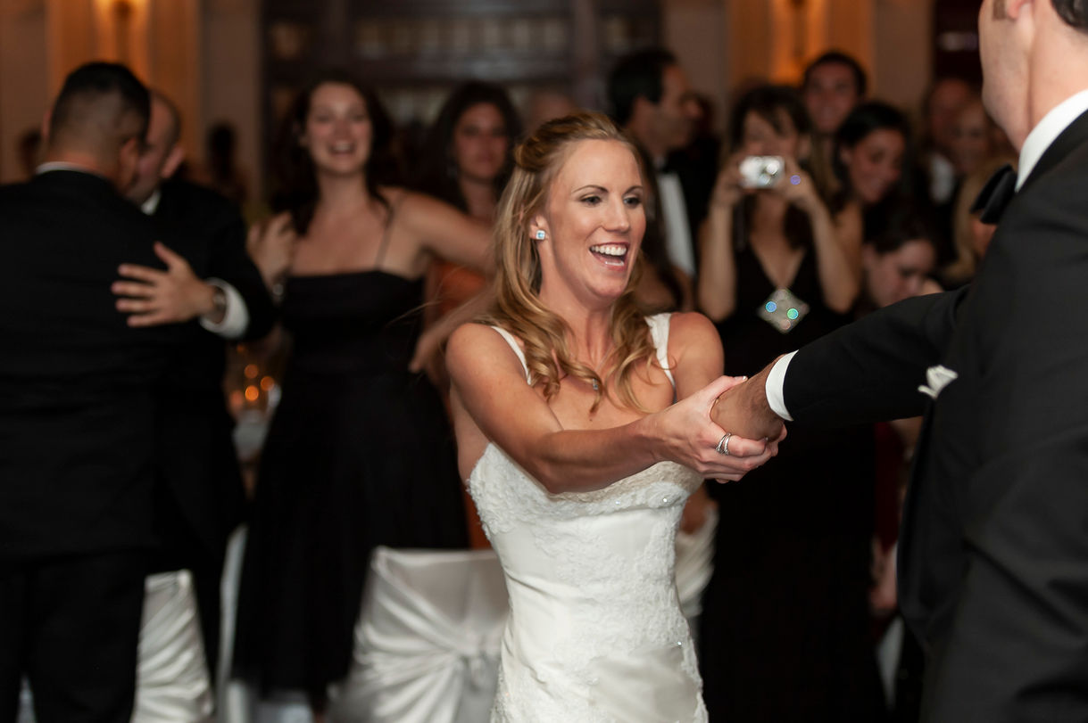The bride and groom share a lively and joyous moment during their first dance. The bride, in a stunning white gown, smiles radiantly as she holds the groom's hand. Guests in the background watch and cheer, capturing the celebratory atmosphere of the reception."
