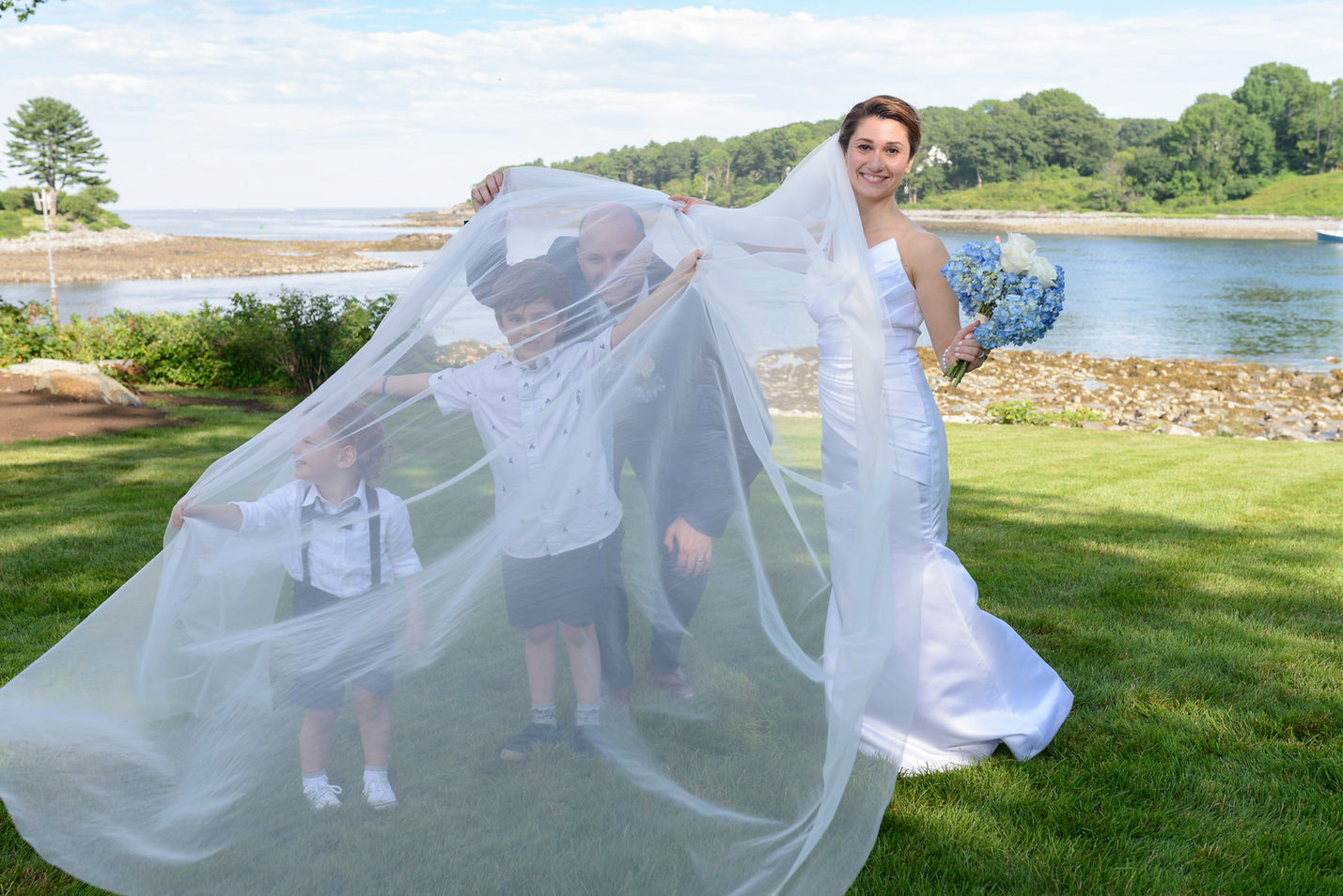 Happy bride holding a bouquet of blue hydrangeas, playfully draping her long veil over two young boys and the groom, with a scenic oceanfront background featuring calm water and lush greenery.
