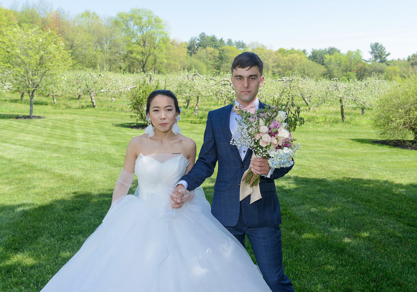 Bride and groom holding hands in an apple blossom orchard under a clear blue sky. The bride wears a voluminous white gown with sheer polka-dot gloves, while the groom is dressed in a navy suit with a pink bow tie, holding a pastel bouquet of roses, hydrangeas, and greenery. The lush orchard creates a serene and romantic backdrop.