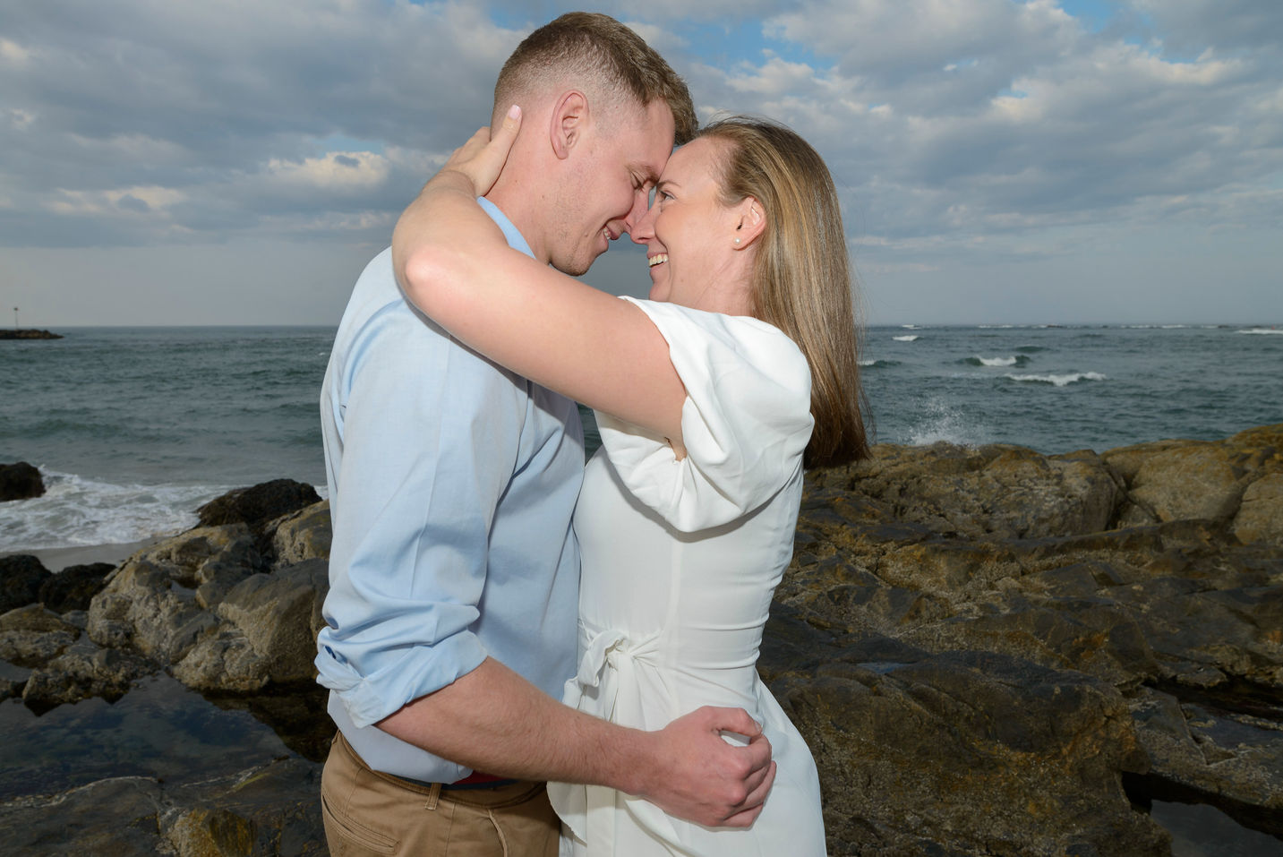 A couple shares an intimate moment during their engagement session, standing on rugged rocks by the ocean. The woman smiles warmly as she holds her partner's face, while the waves crash in the background under a partly cloudy sky. The scene exudes love and connection against the serene coastal setting.