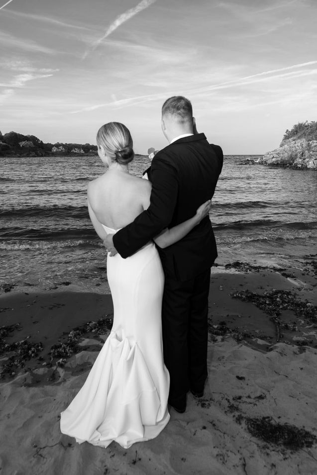 Black and white portrait of the bride and groom embracing on the beach by the water.