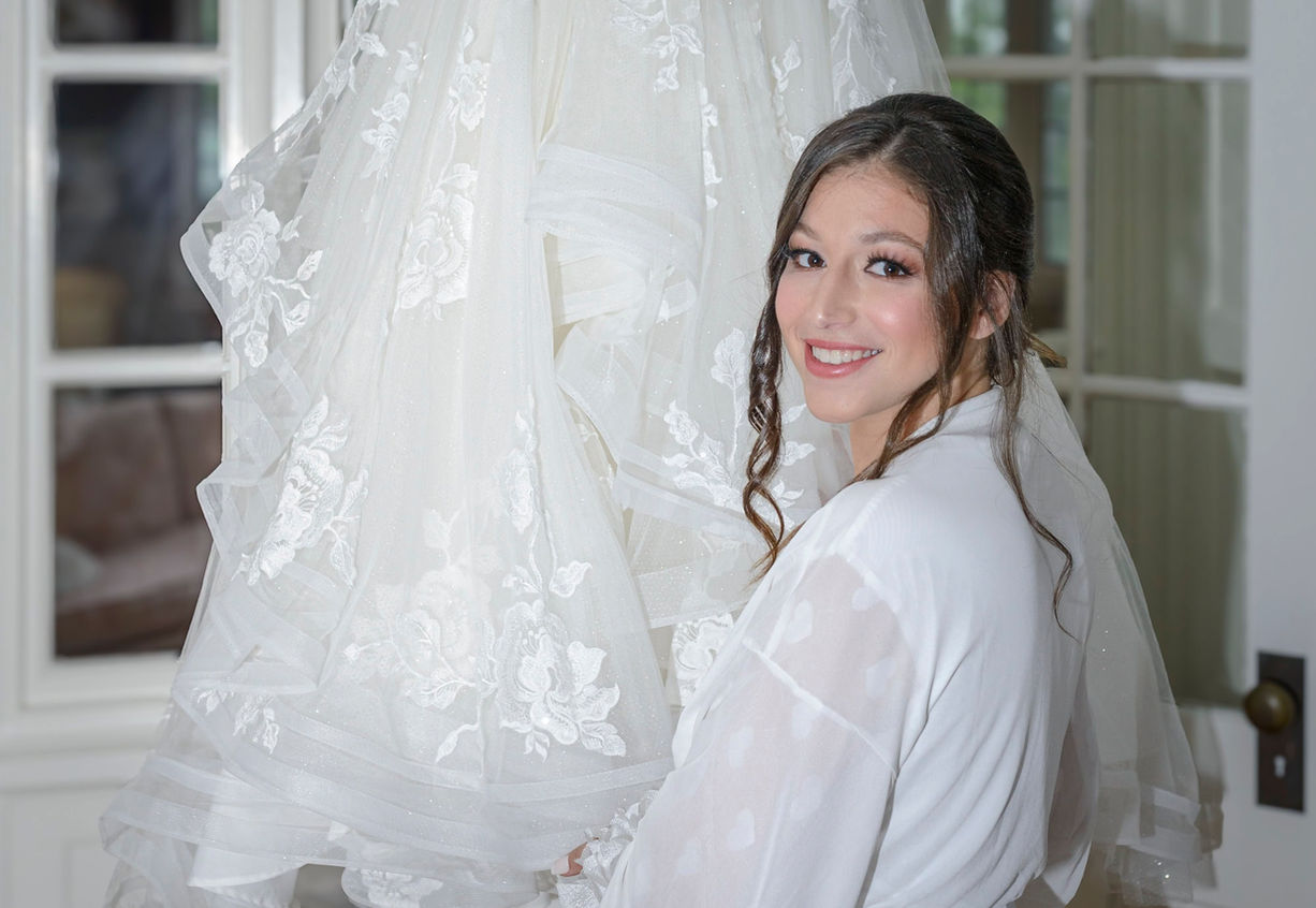 Bride standing next to her white lace wedding dress with white flowers.