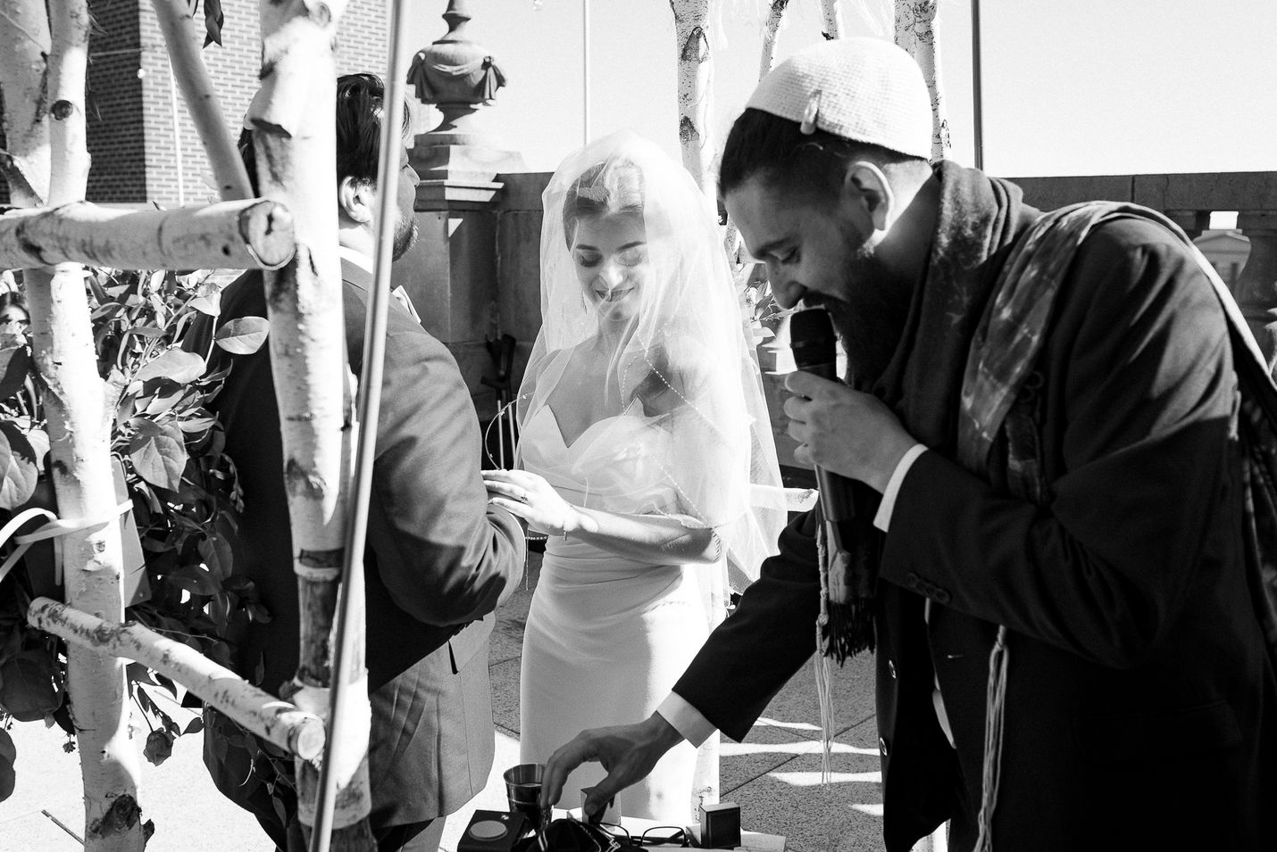 A black-and-white wedding photograph capturing a moment during the rooftop ceremony. The bride, wearing a veil, smiles warmly as she holds the groom's hands under the chuppah. The officiant, holding a microphone, leans forward to adjust ceremonial items on the table. The birchwood chuppah and surrounding decor add a natural and elegant touch to the scene.