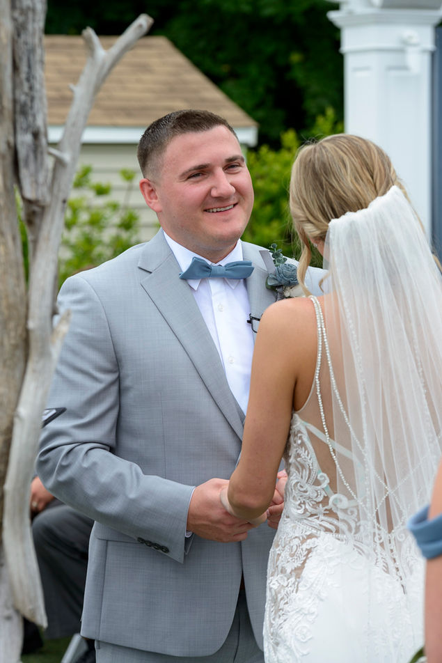 Groom in a light gray suit and blue bowtie smiling at the bride during the wedding ceremony. The bride, seen from the back, wears a lace gown with a long veil flowing down her back.