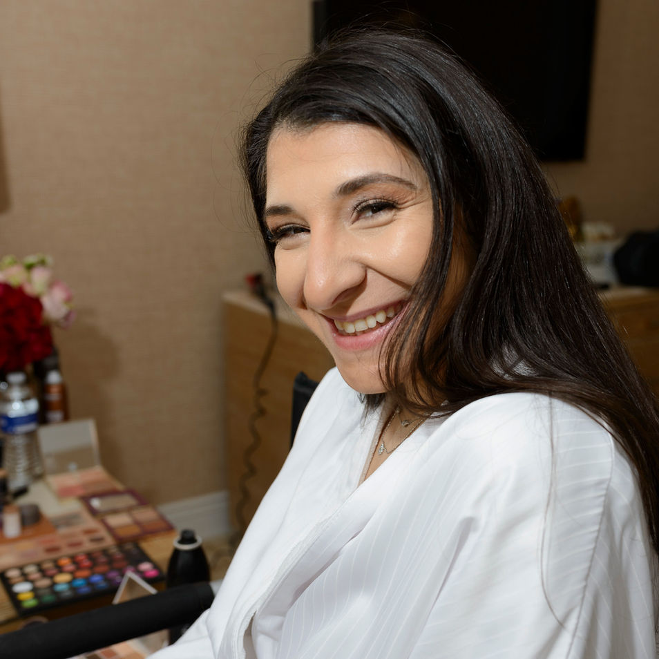 A candid portrait of the bride in her preparation room, flashing a joyful smile while wearing a white robe. The background features a table cluttered with makeup products, adding to the bustling atmosphere of wedding preparations.