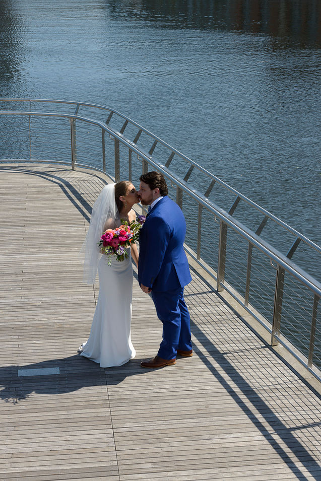 The bride and groom share a tender kiss on a wooden boardwalk by the water, moments after their "First Look." The bride holds a vibrant bouquet of pink, orange, and purple flowers, while her veil flows gently behind her. The groom is dressed in a sharp blue suit, adding a pop of color against the serene waterfront backdrop.