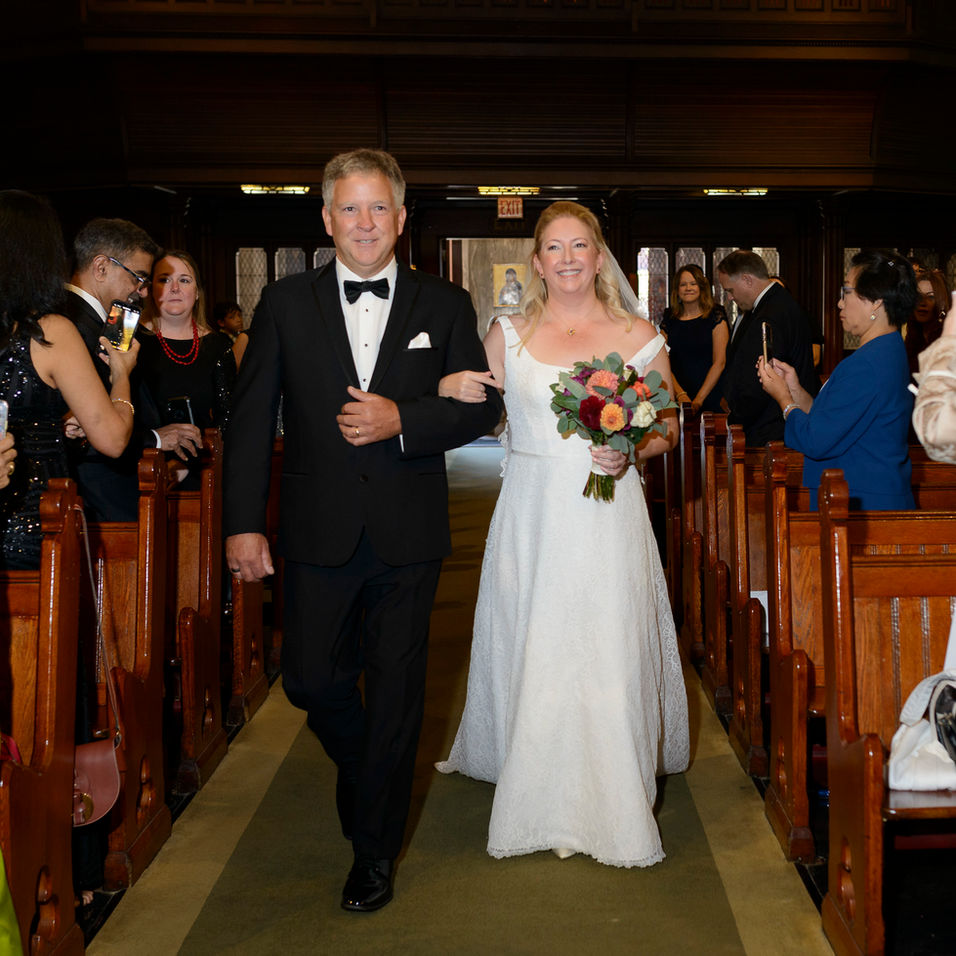A bride walking arm-in-arm with her father down the church aisle. The bride wears a white lace gown and holds a vibrant bouquet of flowers. The father is dressed in a black tuxedo with a bow tie. Guests seated in wooden pews watch and smile as the pair proceeds toward the altar.