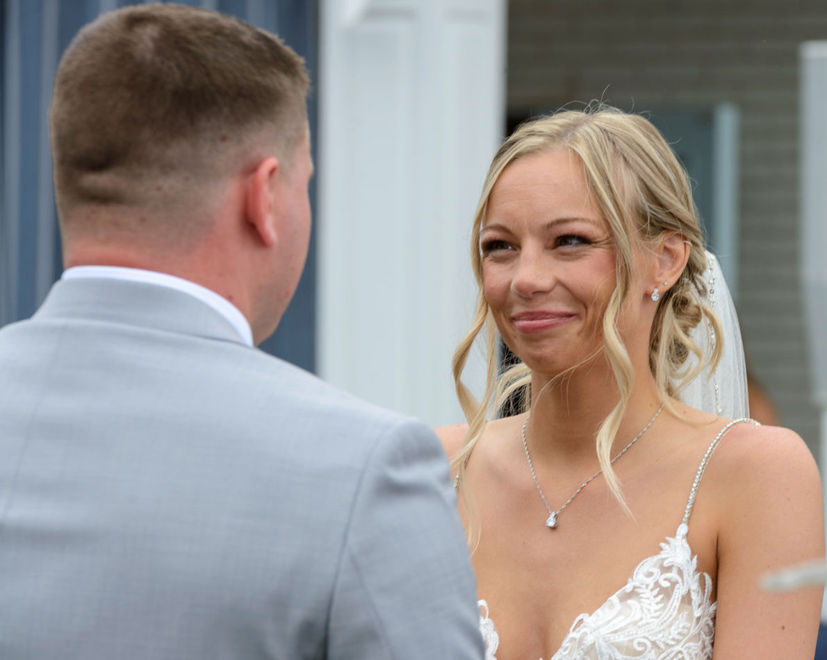 Bride smiling during the wedding ceremony, looking at the groom in a light gray suit. The bride wears a lace wedding gown and delicate necklace, with her hair styled in loose curls and veil flowing behind her.