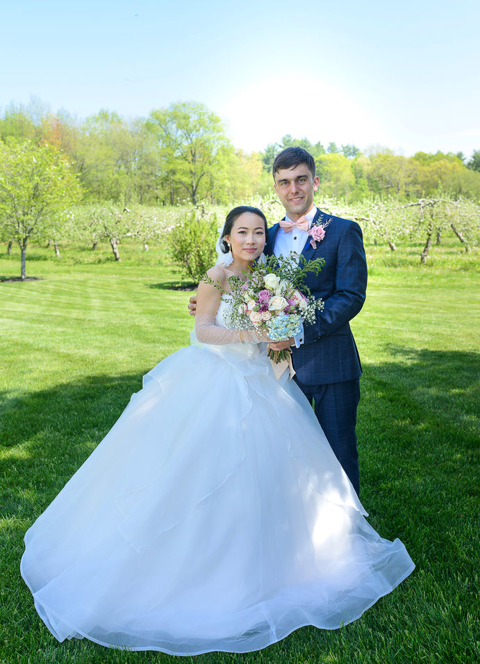 Bride and groom posing together in an apple blossom orchard under a bright blue sky. The bride wears a voluminous white gown and holds a colorful bouquet of roses, hydrangeas, and greenery. The groom, dressed in a navy suit with a pink bow tie and boutonniere, wraps his arm around the bride as they smile warmly at the camera.