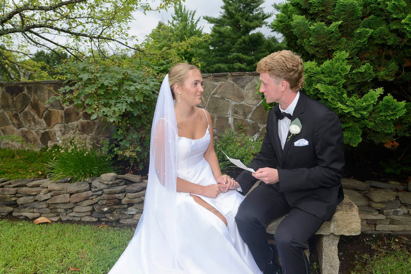 Bride and groom reading vows together on garden bench.