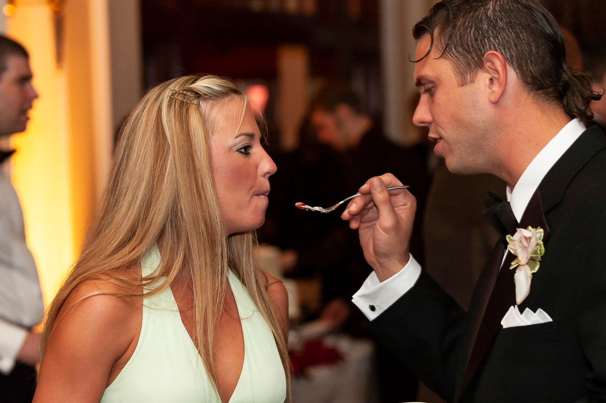 A playful moment during the wedding reception as a male guest in a black tuxedo attempts to feed a piece of cake to his girlfriend. She leans forward with a smile, dressed in a light mint-green dress mouth closed, as the festive ambiance of the celebration surrounds them.