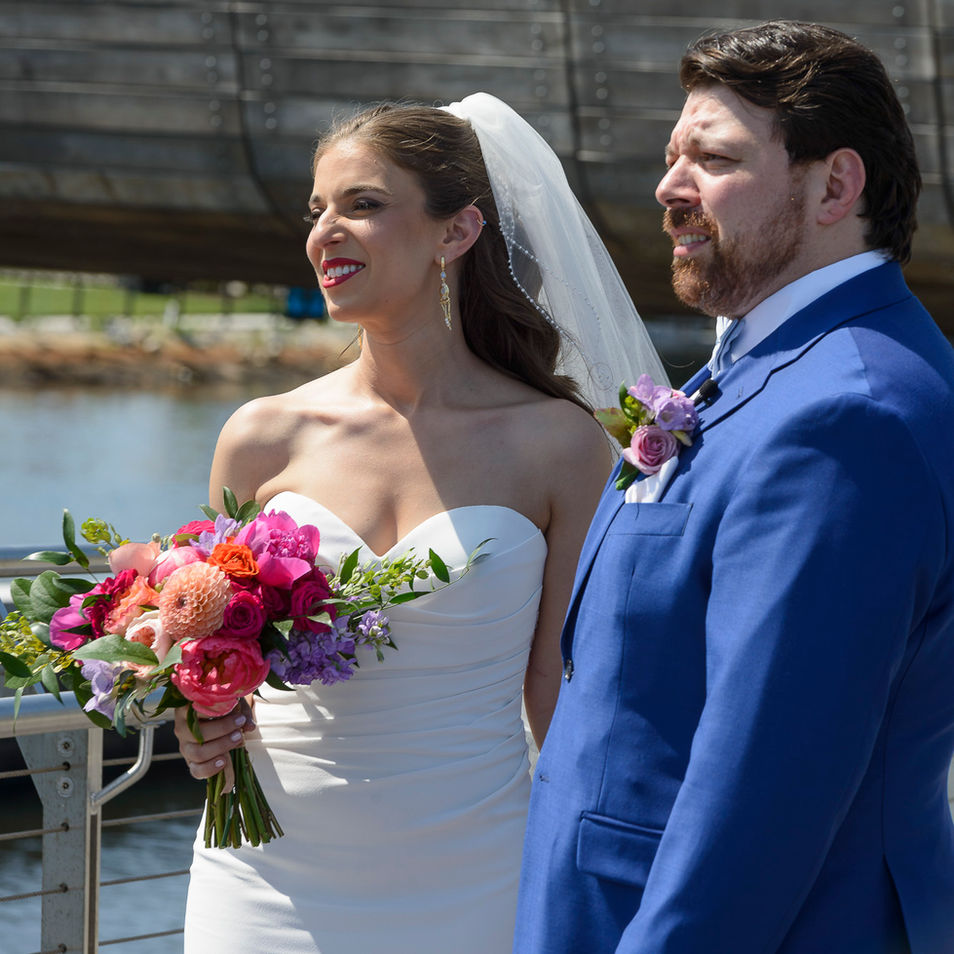 The bride and groom stand side by side on a sunny day, smiling brightly. The bride holds a colorful bouquet of pink, orange, and purple flowers, her veil flowing behind her. The groom is dressed in a vibrant blue suit with a lavender boutonniere, both looking towards the distance with happiness. A calm waterfront and a wooden bridge provide a picturesque backdrop.