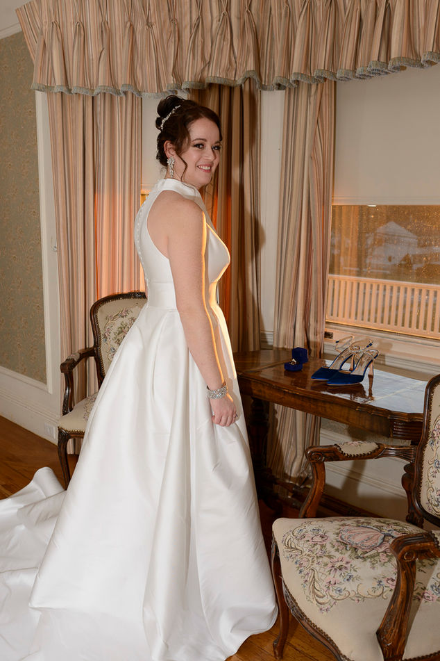 A bride standing in an elegant room, smiling at the camera. She wears a classic white satin wedding gown with a fitted bodice and full skirt, accessorized with diamond earrings and a bracelet. Behind her, a wooden table holds blue high heels and a jewelry box, with vintage chairs and draped curtains completing the setting.