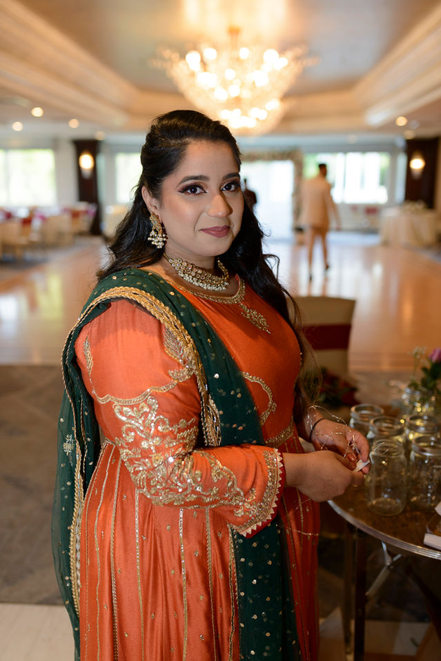A portrait of the bride's sister during a Pakistani wedding reception setup. She is dressed in a traditional red orange outfit adorned with gold embroidery and a green dupatta with intricate embellishments. She stands gracefully beside a table of jars for flowers in an elegant banquet hall with a grand chandelier shining brightly in the background. The setting exudes a festive and welcoming atmosphere.