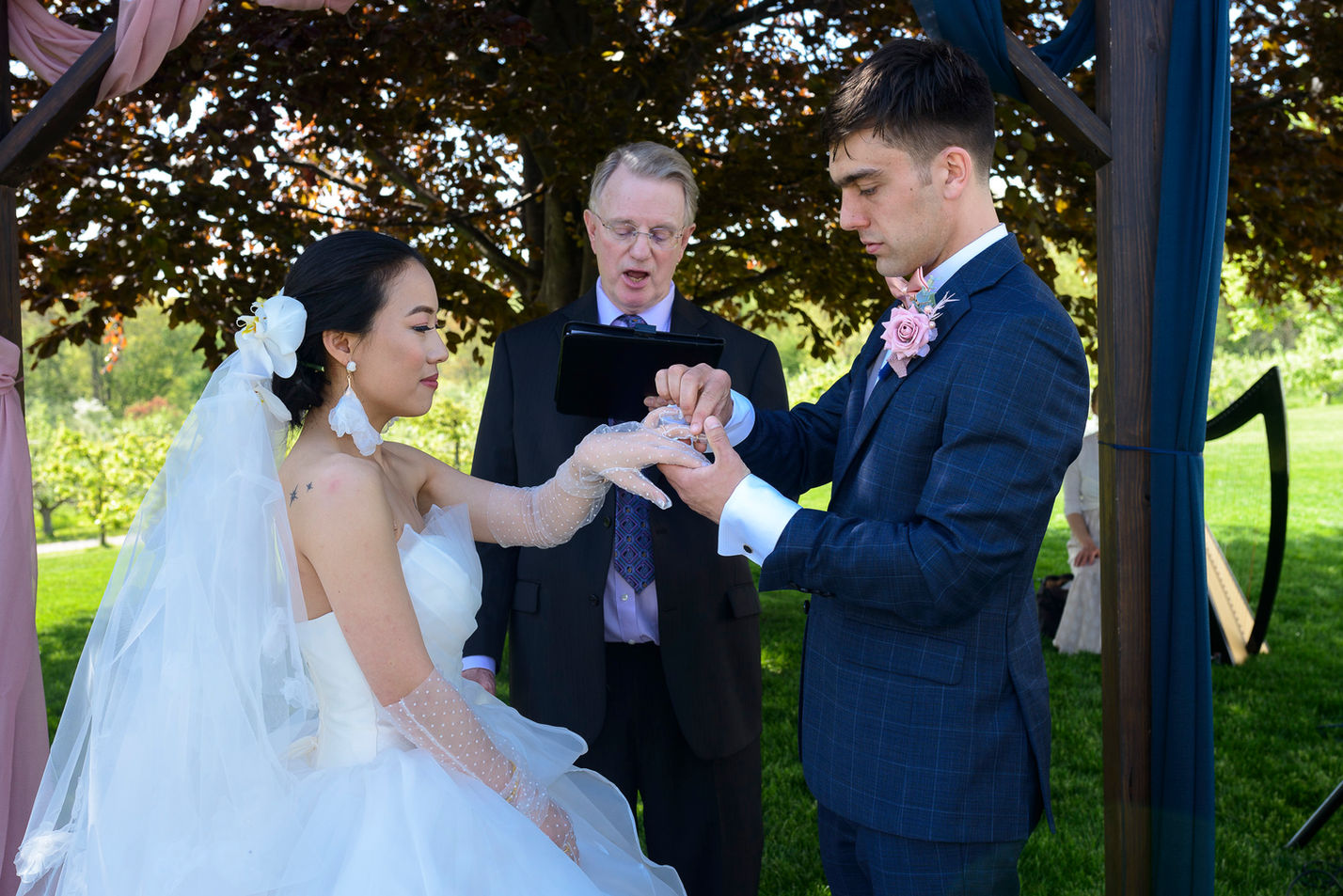 Groom placing a ring on the bride's finger during their wedding ceremony under an arbor. The bride, wearing a white gown with sheer polka-dot gloves, smiles softly, while the officiant stands behind them, reading from a tablet. The outdoor setting features lush greenery and a harp in the background, adding to the romantic atmosphere.