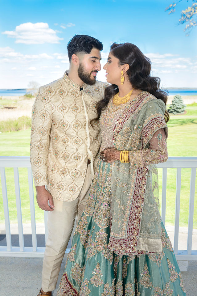 A joyful moment from a Pakistani wedding celebration, featuring a smiling bride and groom in traditional attire. The groom wears a cream-colored sherwani with intricate gold and green embroidery, while the bride dons a stunning sage green and gold lehenga adorned with detailed embellishments. Her outfit is complemented by gold jewelry and traditional mehndi on her hands. They share an affectionate gaze against a backdrop of lush greenery and a bright blue sky with scattered clouds.