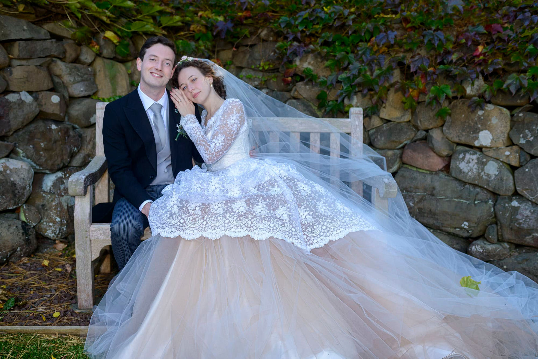 Elegantly dressed wedding couple sitting on a wooden garden bench in front of an old stone wall.