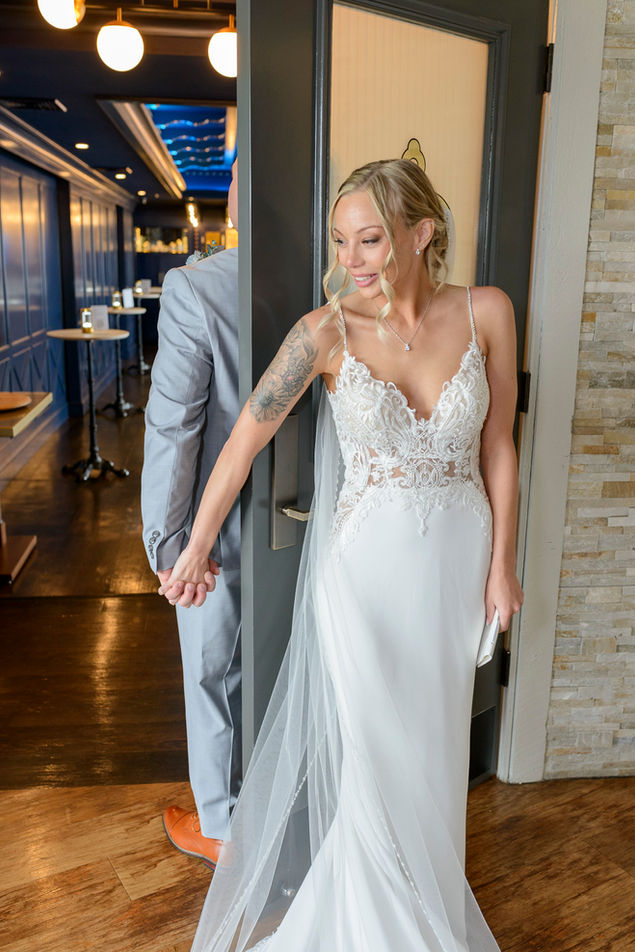 Bride in a white lace wedding gown holding hands with the groom through a doorway, standing apart and unable to see each other before the ceremony. Emotional pre-wedding moment as they prepare to read vows.