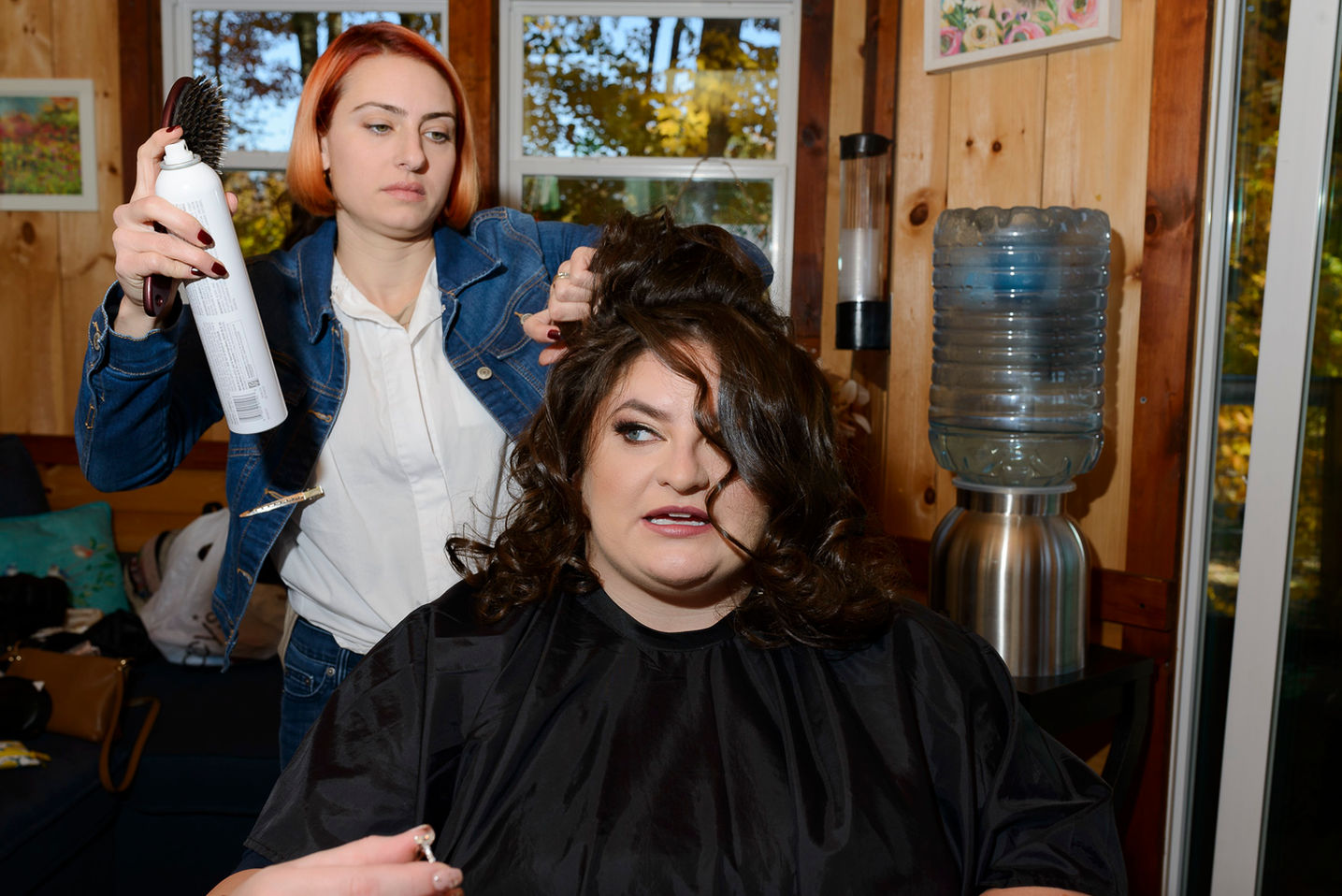 The bride sits in a salon chair as a hairstylist with orange hair applies hairspray while styling her voluminous curls in a warm, rustic room filled with natural light
