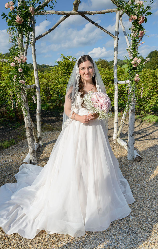 A glowing bride poses under a birchwood arch on her wedding day, wearing a lace gown and a delicate veil edged with floral embroidery. She holds a bouquet of pale pink roses and baby's breath, smiling brightly with sunlight highlighting her joyful expression. The lush vineyard in the background completes the romantic scene.