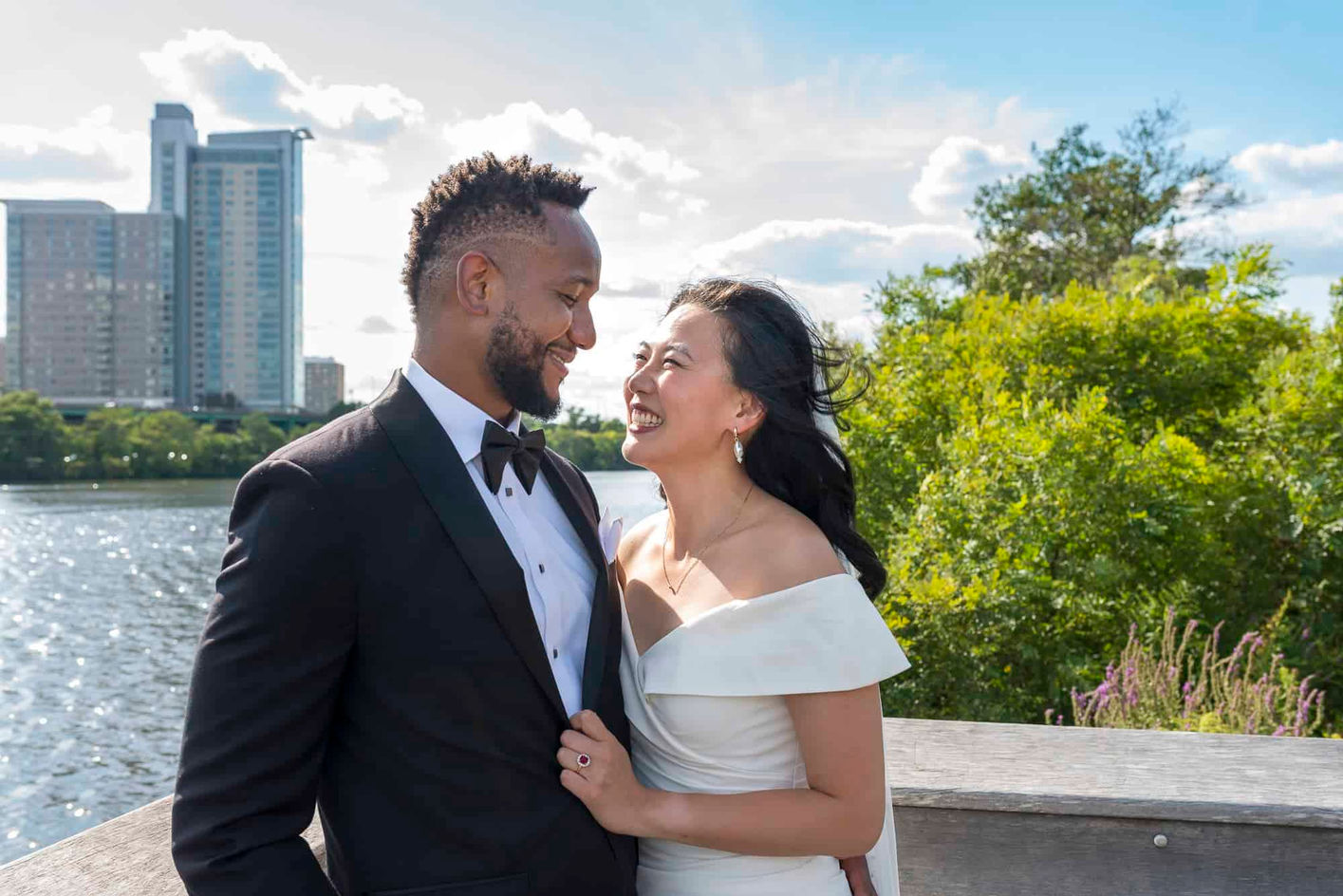 Bride and groom share a joyful moment after their first look on the deck at Magazine Beach in Cambridge, Boston, standing by the Charles River with city buildings and trees in the background.
